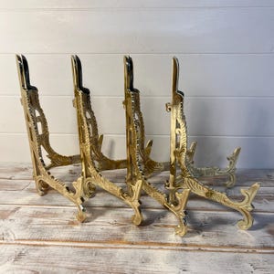 May include: A set of four matching gold-toned metal display stands, each featuring an L-shaped frame with intricate embossed scrollwork patterns along the vertical and horizontal supports, designed to hold items upright on a flat surface.