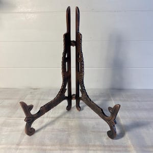 May include: A brown, ornate display stand with a three-legged base and a vertical support. The stand features carved floral designs and is designed to hold plates or artwork. The stand is made of wood and is approximately 12 inches tall.