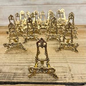 May include: A collection of small, ornate gold-colored display stands. Each stand features a decorative, openwork design with a three-legged base and a top section with a circular opening. The stands are arranged on a wooden surface.