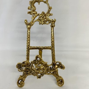 May include: A small, ornate gold-colored easel with intricate floral and scrollwork details. The easel has a decorative top and base, and is approximately 12 inches tall. The design is reminiscent of antique or vintage decor.