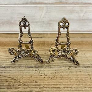 May include: A pair of antique brass display stands. Each stand has an ornate, openwork design with a central support and a base with floral and scroll details. The stands are designed to hold small items.