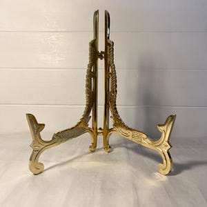 Puede incluir: Un soporte de exhibición de metal dorado con un intrincado patrón en relieve a lo largo de sus dos brazos articulados. El soporte tiene un apoyo vertical central y dos pies curvos que se extienden hacia afuera para sostener objetos en posición vertical.