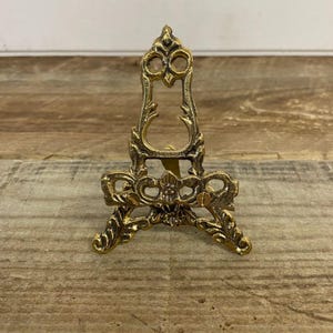 Könnte beinhalten: Verziertes, goldfarbenes Metalleasel mit dekorativem, durchbrochenem Design. Das Staffelei hat einen dreibeinigen Sockel und eine vertikale Rückseite mit gewölbter Oberseite. Die aufwendigen Details deuten auf einen Vintage- oder Antik-Stil hin, geeignet für kleine Kunstwerke.