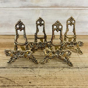 May include: A set of four small, ornate brass display stands. Each stand has a decorative, openwork design with floral and scroll details. The stands are approximately 10 cm tall and are suitable for displaying small items.