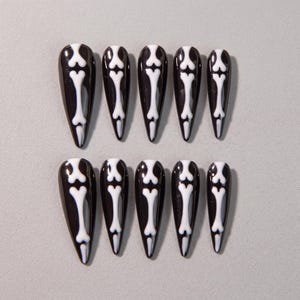 May include: A set of ten press-on nails with a glossy black base and a white bone design running down the centre of each piece. The nails feature a sharp, pointed stiletto shape, arranged in two rows of five, showcasing a high-contrast monochromatic aesthetic.