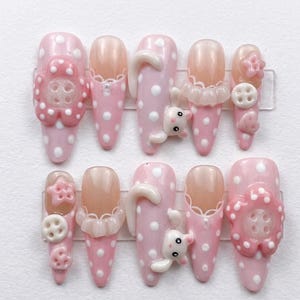 May include: Set of ten pink and white polka dot press-on nails. Some nails feature 3D embellishments, including a white bear, a flower, and a button. The nails have a glossy finish and are arranged on a clear plastic sheet.