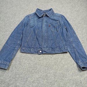 Puede incluir: Chaqueta vaquera azul claro con cierre de botones, cuello y un pequeño bolsillo en el pecho. La chaqueta tiene un diseño clásico con una etiqueta roja en el bolsillo. La chaqueta está tendida sobre una superficie gris.