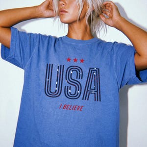 May include: Blue t-shirt with the word "USA" in a bold, outlined font, featuring red and white stripes and stars. Above the word are three red stars, and below it, the phrase "I BELIEVE" in red. The shirt has a vintage, faded look.