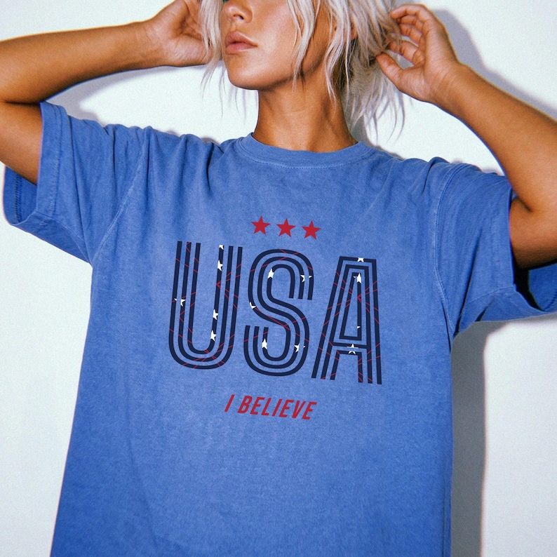 May include: Blue t-shirt with the word "USA" in a bold, outlined font, featuring red and white stripes and stars. Above the word are three red stars, and below it, the phrase "I BELIEVE" in red. The shirt has a vintage, faded look.