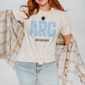 May include: Cream-colored t-shirt with the word "ARG" in light blue, with a sunburst graphic above and the word "MUCHACHOS" below. The shirt is knotted at the waist. A plaid shirt is draped over the shoulders.