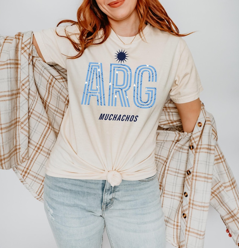 May include: Cream-colored t-shirt with the word "ARG" in light blue, with a sunburst graphic above and the word "MUCHACHOS" below. The shirt is knotted at the waist. A plaid shirt is draped over the shoulders.