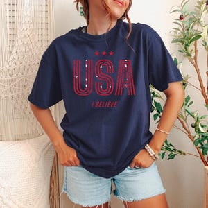 May include: Navy blue t-shirt with the word "USA" in red, distressed lettering, and the phrase "I BELIEVE" below. The shirt also features three red stars above the word "USA".