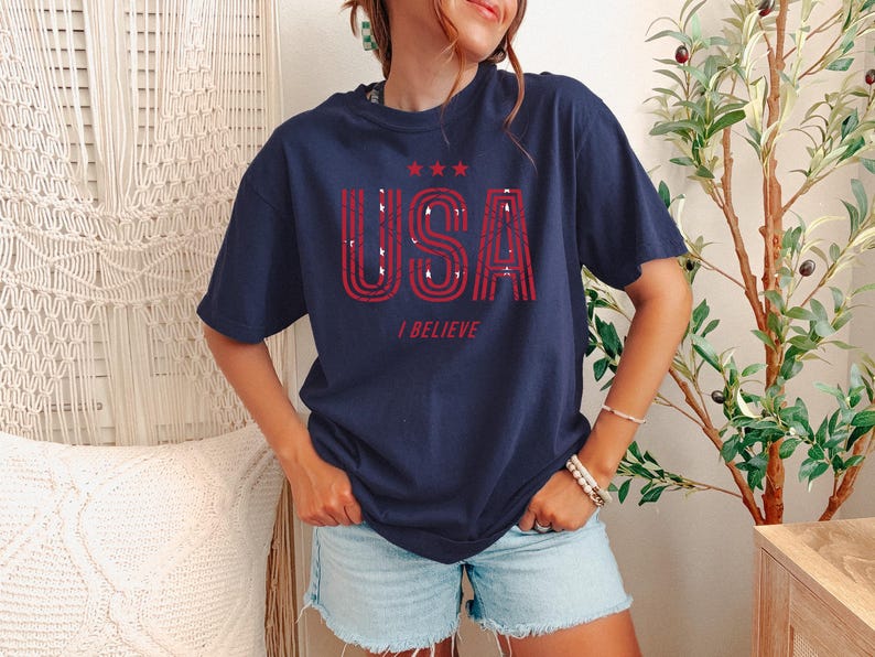 May include: Navy blue t-shirt with the word "USA" in red, distressed lettering, and the phrase "I BELIEVE" below. The shirt also features three red stars above the word "USA".