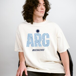 May include: Cream-colored t-shirt with the word "ARG" in blue, striped letters, and the word "MUCHACHOS" below. A blue sunburst graphic is above the "ARG". The shirt is worn by a person sitting on a chair.