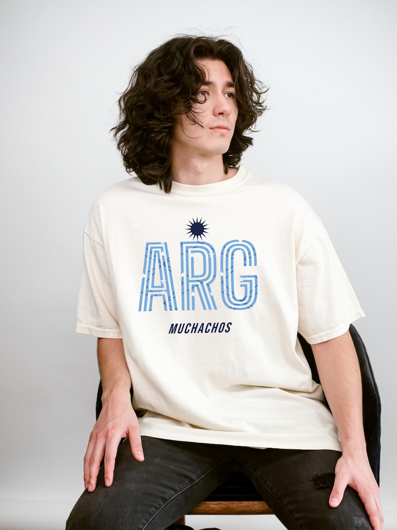 May include: Cream-colored t-shirt with the word "ARG" in blue, striped letters, and the word "MUCHACHOS" below. A blue sunburst graphic is above the "ARG". The shirt is worn by a person sitting on a chair.