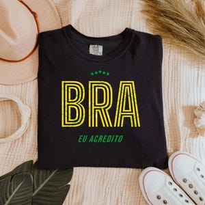 May include: Black t-shirt with the word "BRA" in yellow and green, with the text "EU ACREDITO" below. The shirt is surrounded by a beige hat, white headband, scrunchie, leaves, and white sneakers.