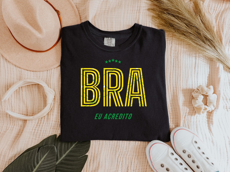 May include: Black t-shirt with the word "BRA" in yellow and green, with the text "EU ACREDITO" below. The shirt is surrounded by a beige hat, white headband, scrunchie, leaves, and white sneakers.