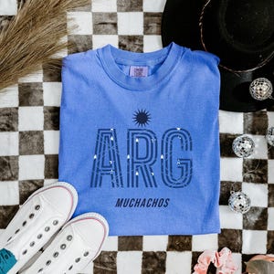 May include: A light blue t-shirt with the word "ARG" in a stylized font, with the word "MUCHACHOS" below. The shirt is laid on a black and white checkered surface, with white sneakers and disco balls.