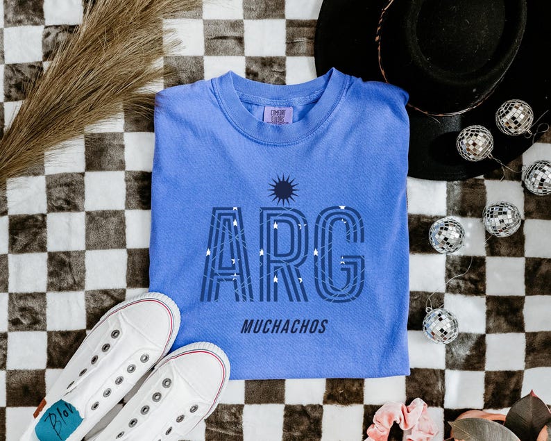 May include: A light blue t-shirt with the word "ARG" in a stylized font, with the word "MUCHACHOS" below. The shirt is laid on a black and white checkered surface, with white sneakers and disco balls.