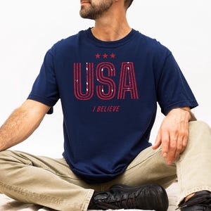 May include: Navy blue t-shirt with the word "USA" in red, outlined letters and three red stars above it. Below the word "USA" is the text "I BELIEVE". The shirt is worn by a person sitting cross-legged.