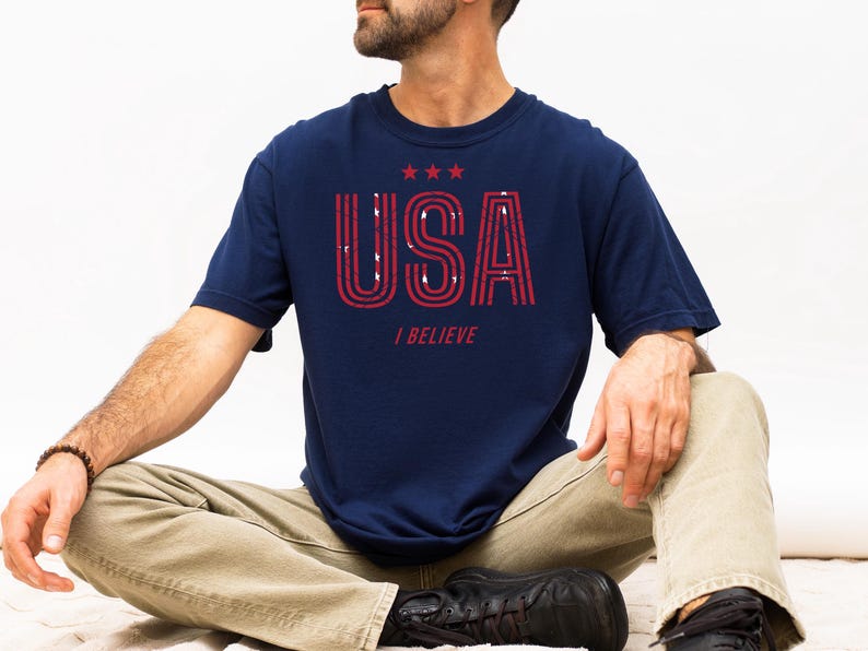 May include: Navy blue t-shirt with the word "USA" in red, outlined letters and three red stars above it. Below the word "USA" is the text "I BELIEVE". The shirt is worn by a person sitting cross-legged.