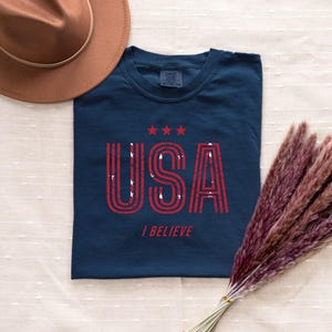 May include: Navy blue t-shirt with the word "USA" in red, distressed lettering, and three red stars above it. The phrase "I BELIEVE" is printed below. A brown hat and a bundle of dried, reddish-brown plants are also in the image.