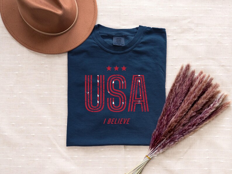 May include: Navy blue t-shirt with the word "USA" in red, distressed lettering, and three red stars above it. The phrase "I BELIEVE" is printed below. A brown hat and a bundle of dried, reddish-brown plants are also in the image.