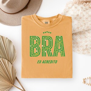 May include: Mustard-colored t-shirt with the word "BRA" in green, outlined with a striped design. The text "EU ACREDITO" is below the word "BRA". The shirt has a crew neck and short sleeves.