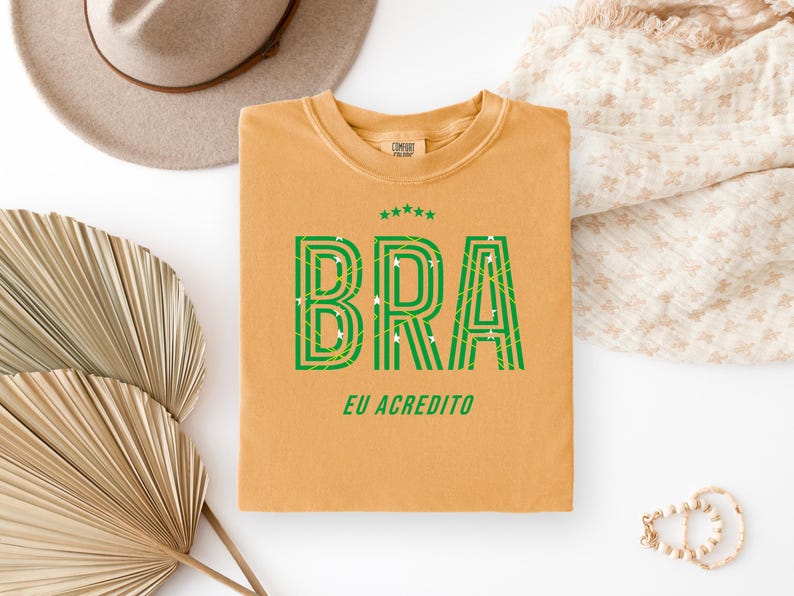 May include: Mustard-colored t-shirt with the word "BRA" in green, outlined with a striped design. The text "EU ACREDITO" is below the word "BRA". The shirt has a crew neck and short sleeves.