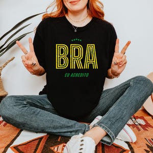 May include: Black t-shirt with the word "BRA" in yellow outlined letters and the text "EU ACREDITO" in green. The shirt is worn with blue jeans. A straw hat and a potted plant are in the background.