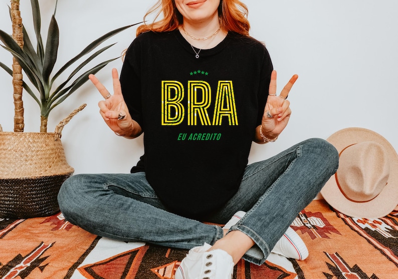 May include: Black t-shirt with the word "BRA" in yellow outlined letters and the text "EU ACREDITO" in green. The shirt is worn with blue jeans. A straw hat and a potted plant are in the background.