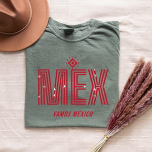 May include: Sage green t-shirt with the word "MEX" in red, stylized with vertical lines and star accents. Below, the text reads "VAMOS M&Eacute;XICO." A brown hat and dried flowers are also in the image.
