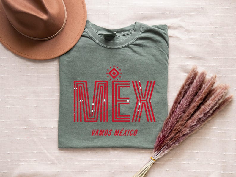 May include: Sage green t-shirt with the word "MEX" in red, stylized with vertical lines and star accents. Below, the text reads "VAMOS M&Eacute;XICO." A brown hat and dried flowers are also in the image.
