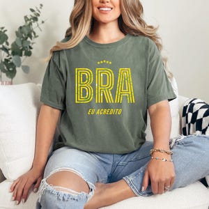 May include: Olive green t-shirt with the word "BRA" in yellow, outlined in a striped design, and the text "EU ACREDITO" below. The shirt is worn with blue jeans. The overall theme is casual wear.