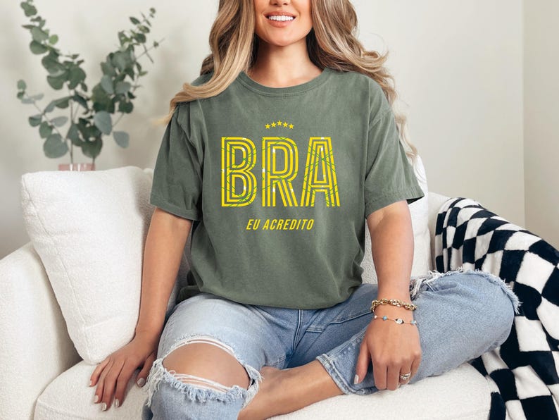 May include: Olive green t-shirt with the word "BRA" in yellow, outlined in a striped design, and the text "EU ACREDITO" below. The shirt is worn with blue jeans. The overall theme is casual wear.