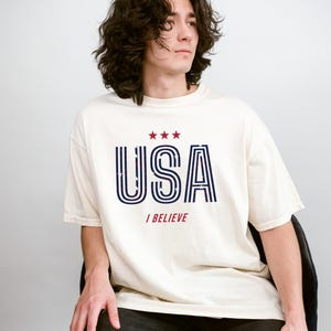 May include: Cream-colored t-shirt with the word "USA" in navy blue and red, with three red stars above the text. The phrase "I BELIEVE" is printed below the word "USA". The shirt is worn by a person.