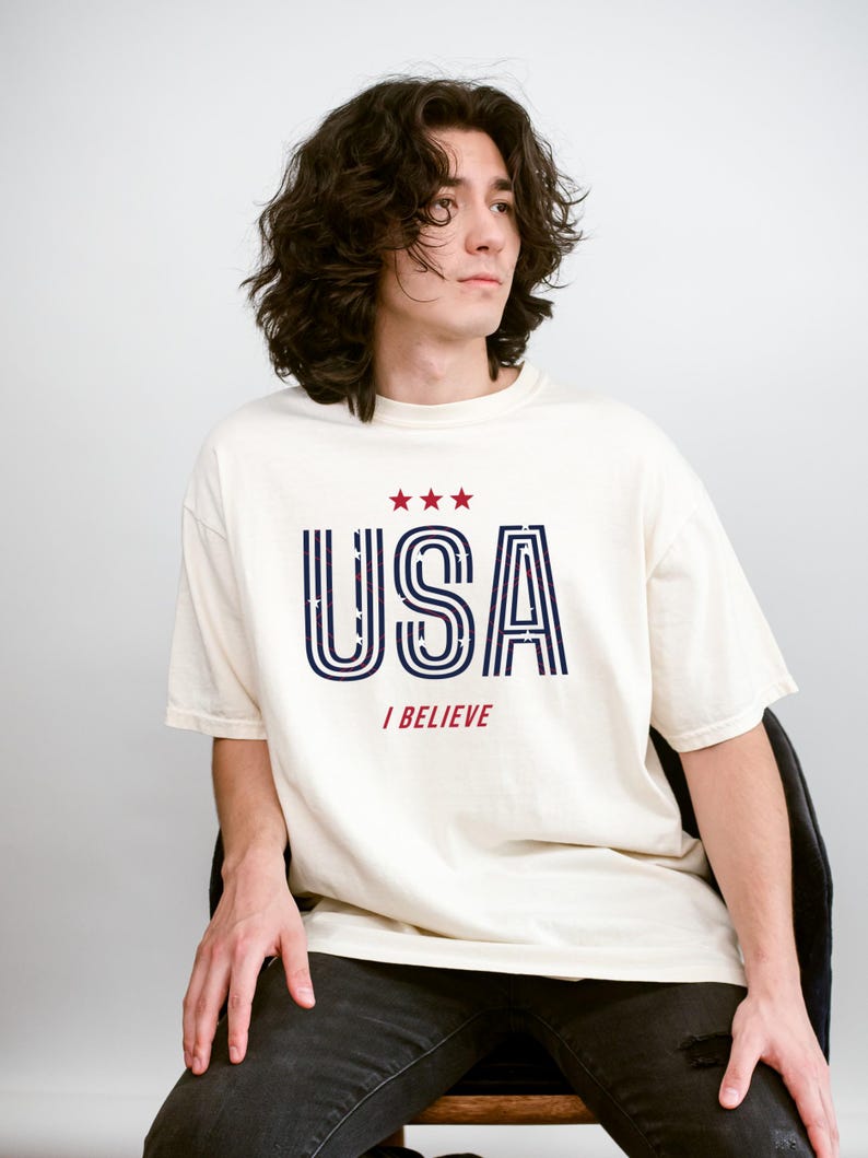 May include: Cream-colored t-shirt with the word "USA" in navy blue and red, with three red stars above the text. The phrase "I BELIEVE" is printed below the word "USA". The shirt is worn by a person.
