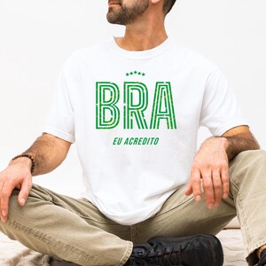 May include: White t-shirt with the word "BRA" in green, outlined with a darker green, and the text "EU ACREDITO" below. The shirt has a crew neck and short sleeves. The person is wearing khaki pants and black shoes.