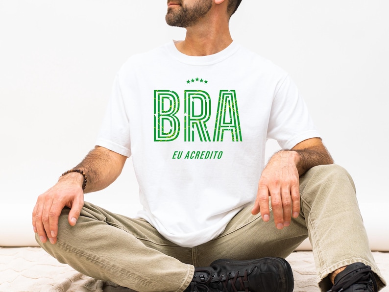 May include: White t-shirt with the word "BRA" in green, outlined with a darker green, and the text "EU ACREDITO" below. The shirt has a crew neck and short sleeves. The person is wearing khaki pants and black shoes.