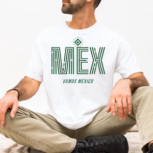 May include: White t-shirt with the word "MEX" in green, featuring a geometric design. Below the word "MEX" is the phrase "VAMOS M&Eacute;XICO" in green. The shirt is worn by a person sitting cross-legged.