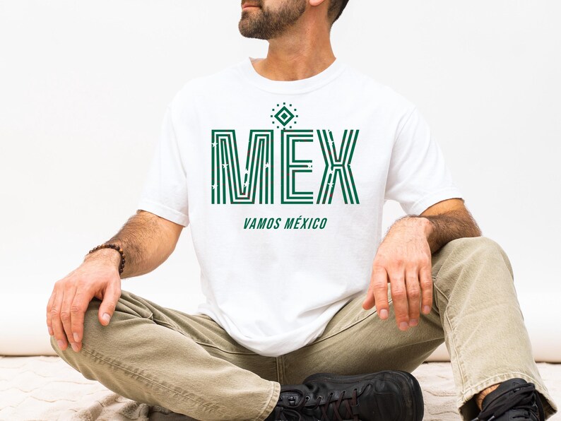 May include: White t-shirt with the word "MEX" in green, featuring a geometric design. Below the word "MEX" is the phrase "VAMOS M&Eacute;XICO" in green. The shirt is worn by a person sitting cross-legged.