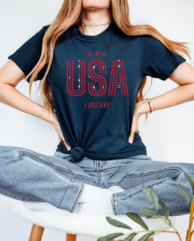May include: Navy blue t-shirt with the word "USA" in red, outlined with white, and the phrase "I BELIEVE" below. The shirt has three red stars above the word "USA".