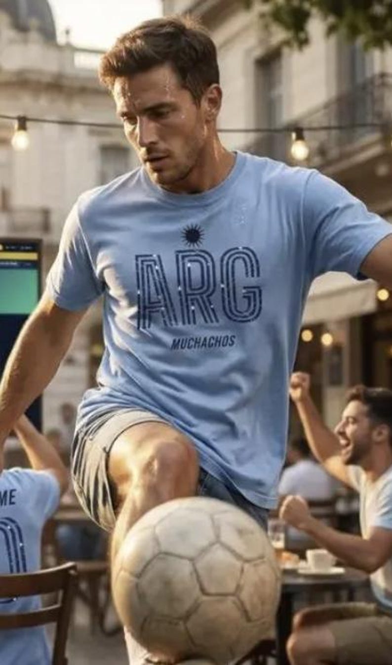 May include: A light blue t-shirt with the word "ARG" in a stylized font, with the word "Muchachos" below it. The shirt is worn by a person playing soccer with a soccer ball. The person is wearing gray shorts.