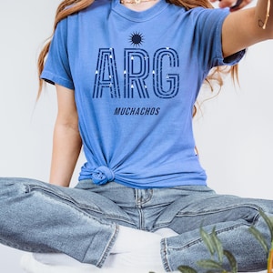 May include: A light blue t-shirt with the word "ARG" in a navy blue font, with a sun graphic above and the word "MUCHACHOS" below. The shirt is knotted at the waist. The person is wearing blue jeans and white socks.