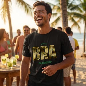 May include: A black t-shirt with the word "BRA" in yellow and green, with the phrase "EU ACREDITO" below. The shirt is worn by a smiling person on a beach, with a group of people and cocktails in the background.
