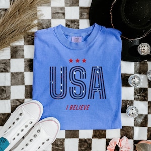 May include: A light blue t-shirt with the word "USA" in a retro font, with three red stars above and the words "I BELIEVE" below. White sneakers and disco balls are also in the image.