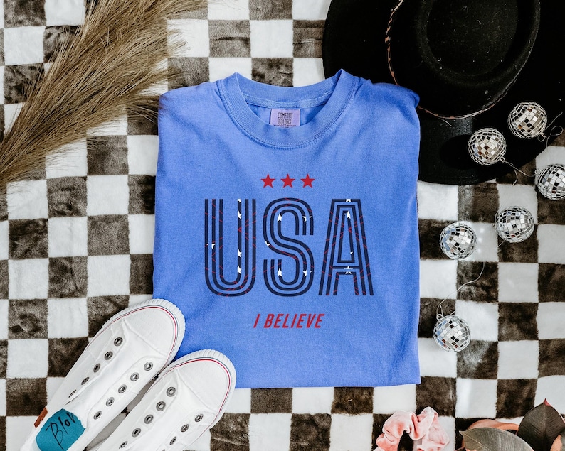 May include: A light blue t-shirt with the word "USA" in a retro font, with three red stars above and the words "I BELIEVE" below. White sneakers and disco balls are also in the image.