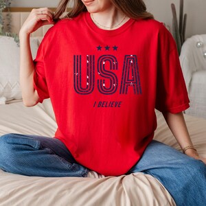 May include: A red t-shirt with the word "USA" in navy blue, along with three stars and the phrase "I BELIEVE". The shirt is worn with blue jeans. The overall theme is patriotic.