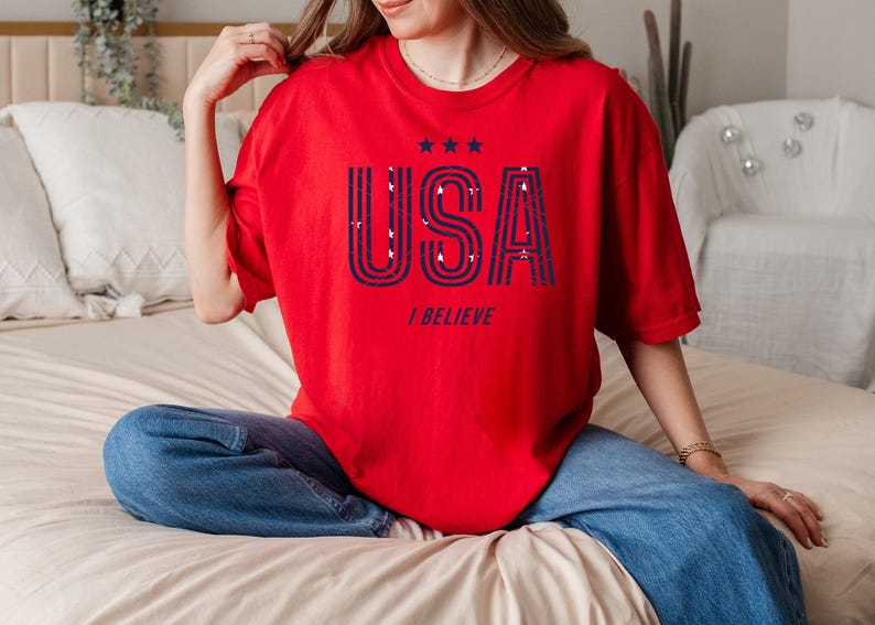 May include: A red t-shirt with the word "USA" in navy blue, along with three stars and the phrase "I BELIEVE". The shirt is worn with blue jeans. The overall theme is patriotic.