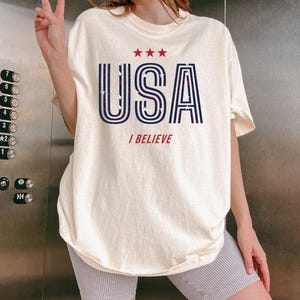 May include: Cream-colored t-shirt with the word "USA" in navy blue, outlined in white, and filled with a star pattern. Above the word are three red stars, and below it, the words "I BELIEVE" in red.